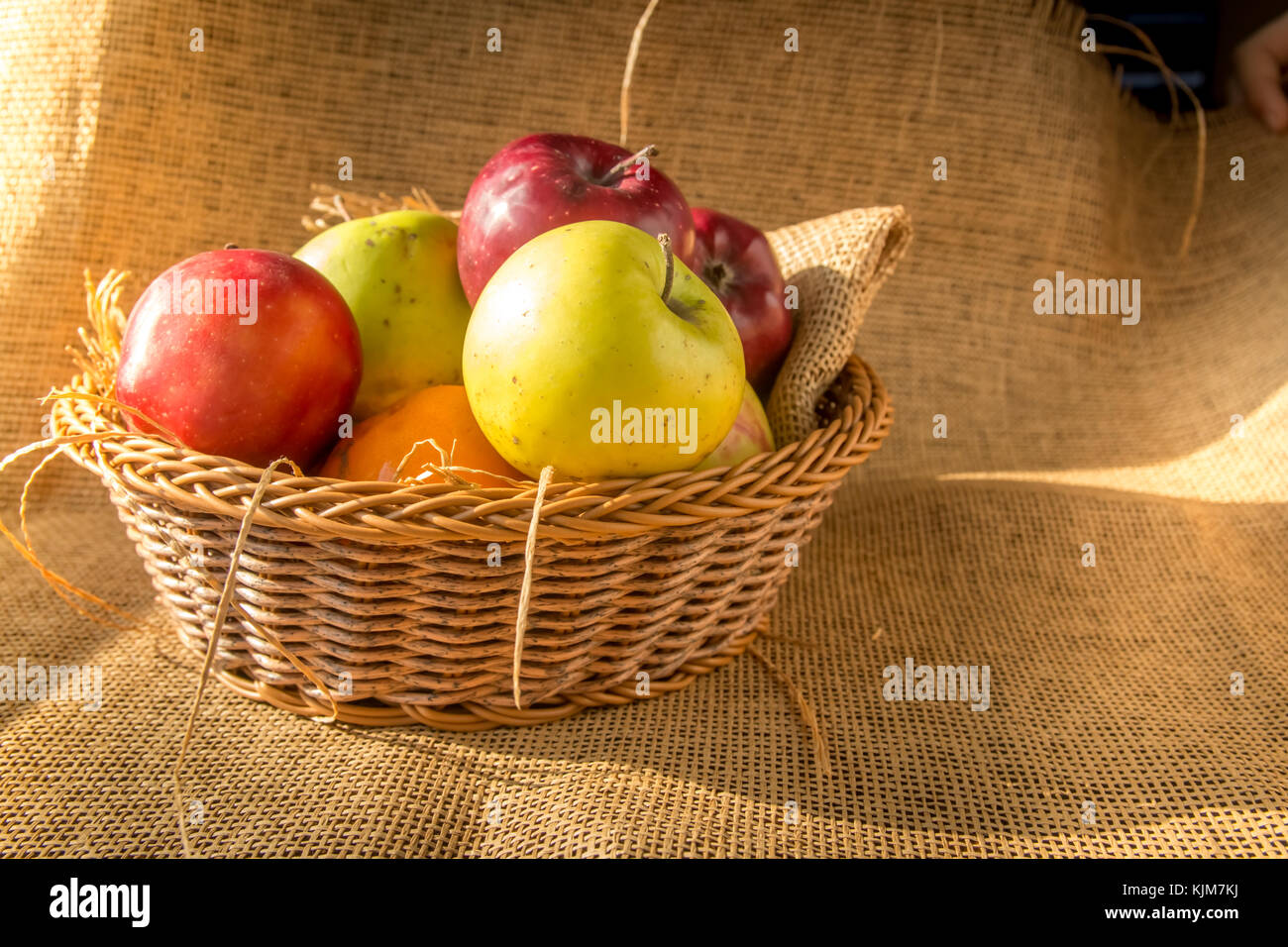 Green and red apples in a basket on burlap background with morning sun shine. Stock Photo