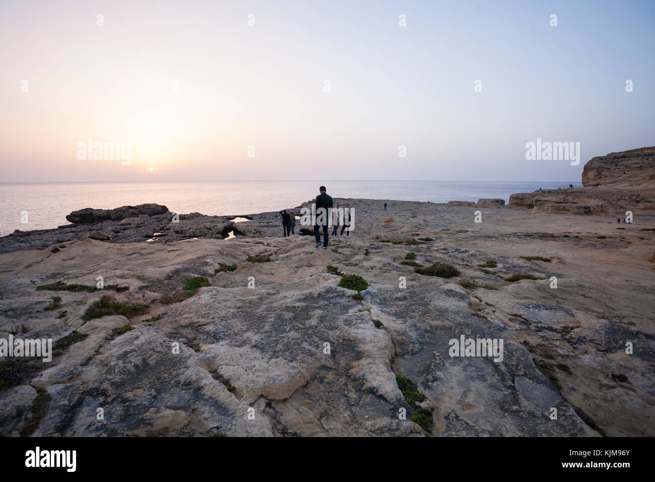 People walking at the sunset hour at Dwejra Bay, looking at sea, with rocky shore foreground. Stock Photo