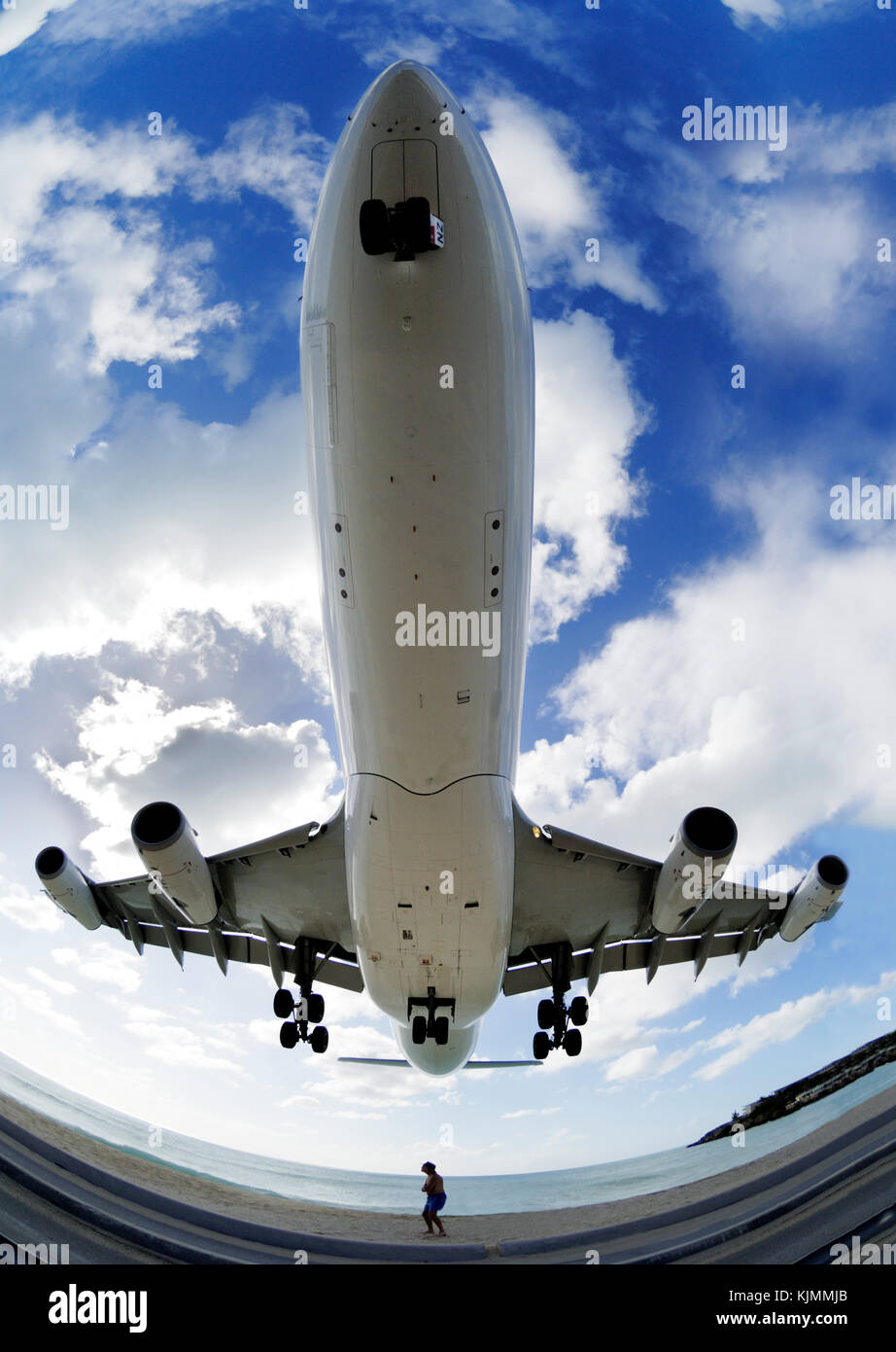 a man in a hat cowering as an A340 passes overhead on low final-approach landing over the sea at Maho Beach with clouds Stock Photo