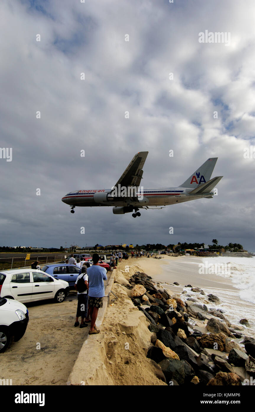 low final-approach landing over Maho Beach on a grey-cloudy-day with cars, rocks, sea and surf splashing the in foreground Stock Photo