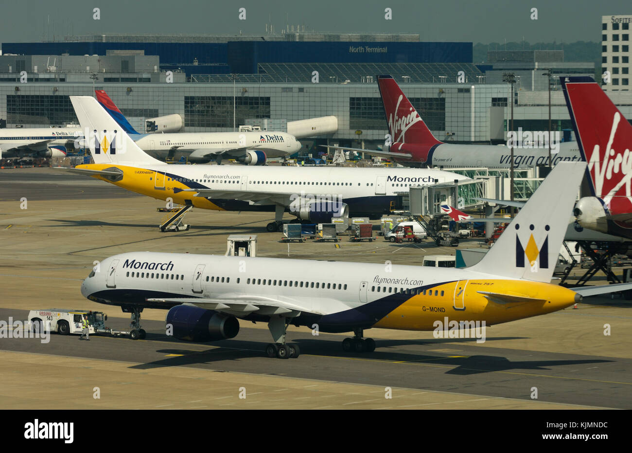 a Monarch Boeing 757-200 being towed by a tug with an Airbus A300, two Virgin Atlantic 747-400s and Delta Air Lines 767-300 parked at the terminal and Stock Photo