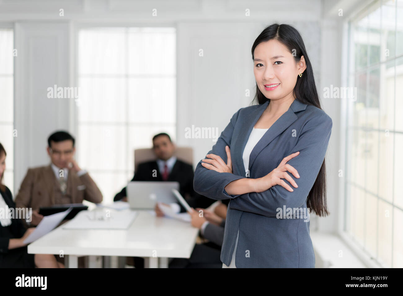 Happy businesswoman standing with arms crossed at office while colleagues discussing in background. Stock Photo