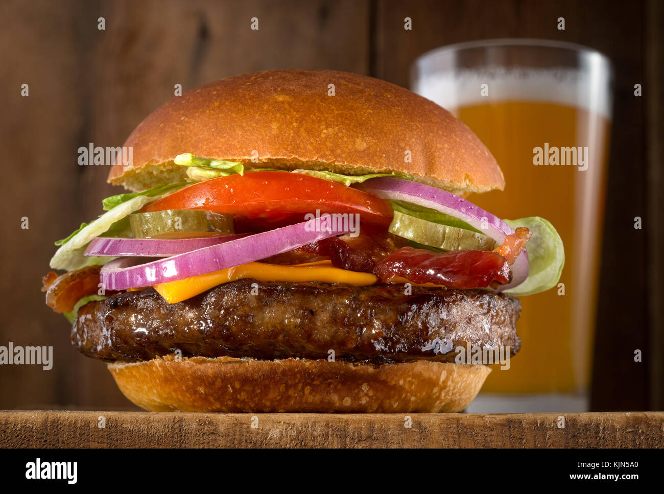 A juicy delicious cheeseburger with bacon, lettuce, tomato, red onions and pickle with a glass of beer. Stock Photo