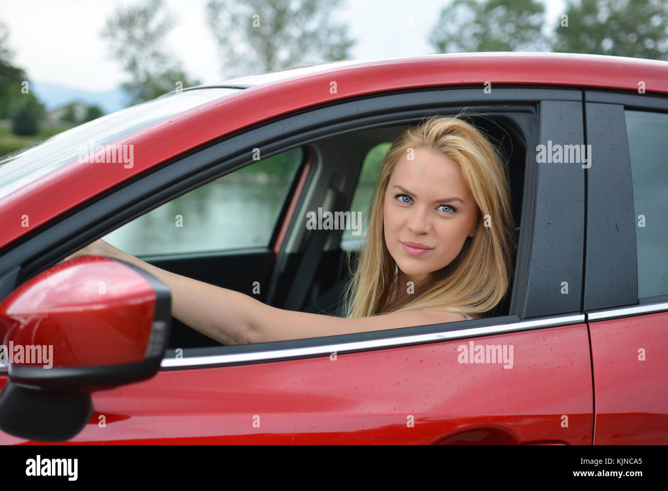 Beautiful young blonde girl driving slowly and carefully brand new red car and looking through open window Stock Photo