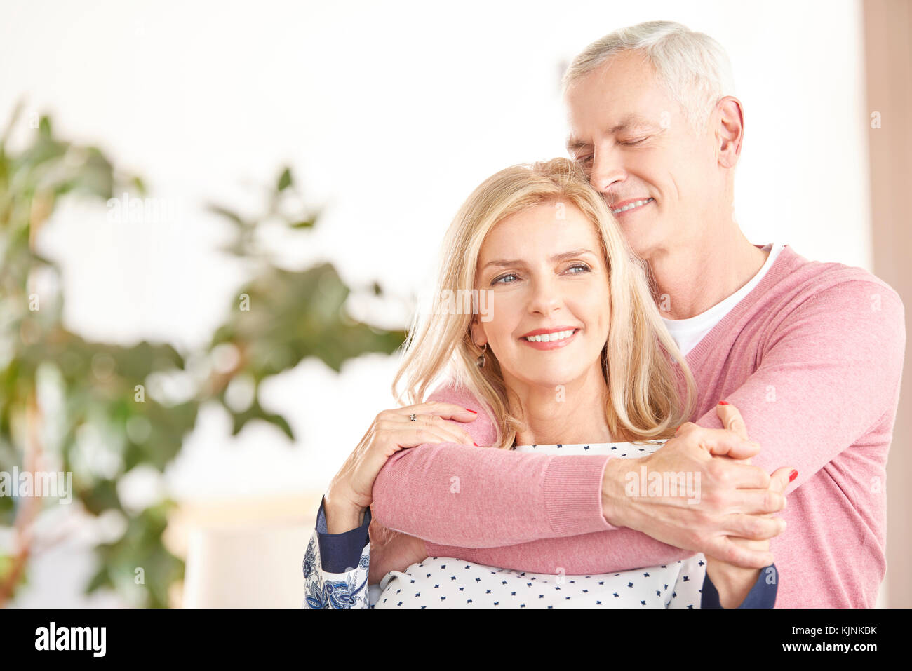 Shot of a happy senior couple standing together. Elderly man hugging her wife from behind while at home in living room. Stock Photo