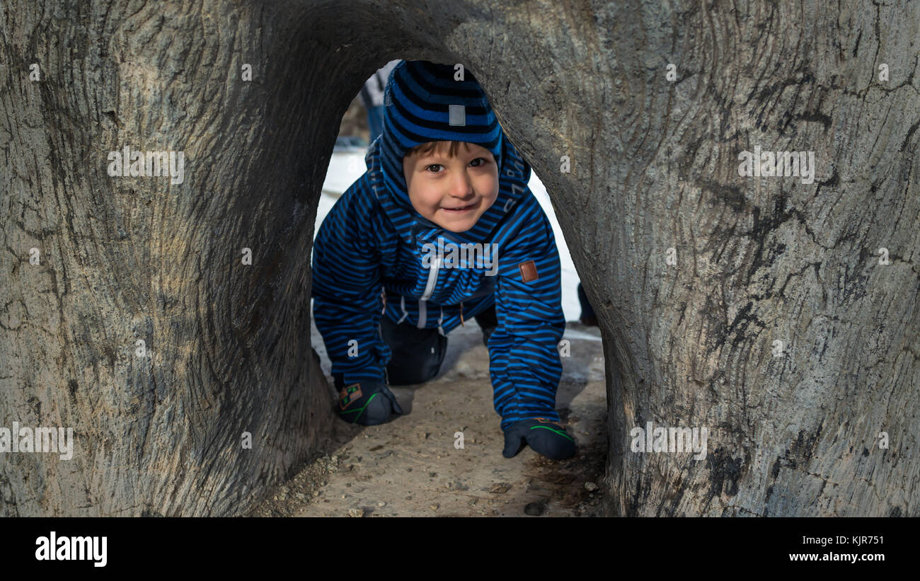 Beautiful kid in warm clothes and hat playing and crawls under the tree in winter time Stock Photo