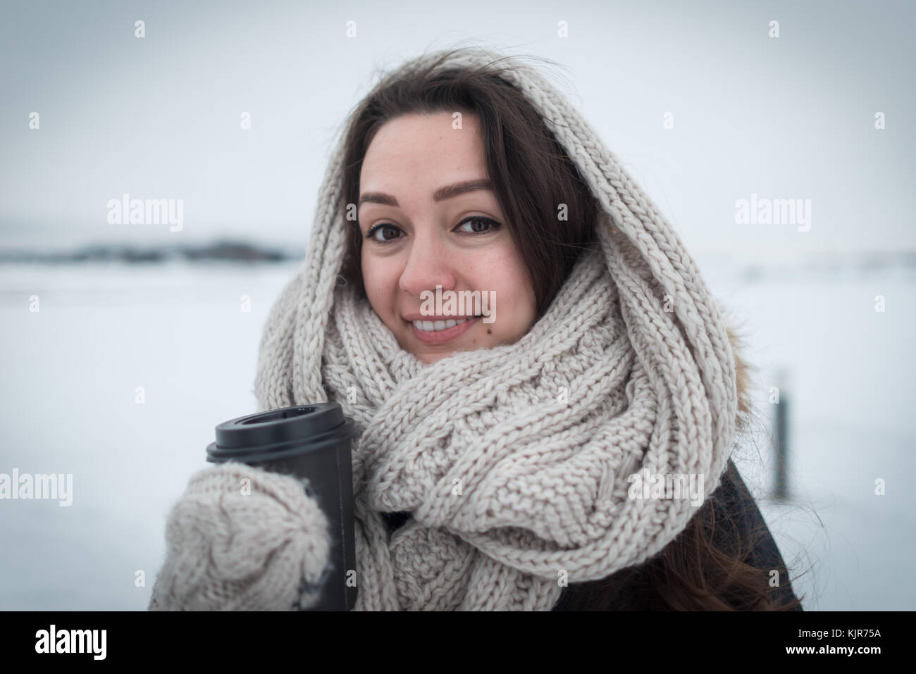 Young woman in warm and soft scarf with hot coffee in cup for take away staying at clear field on the winter background. Cold day Stock Photo