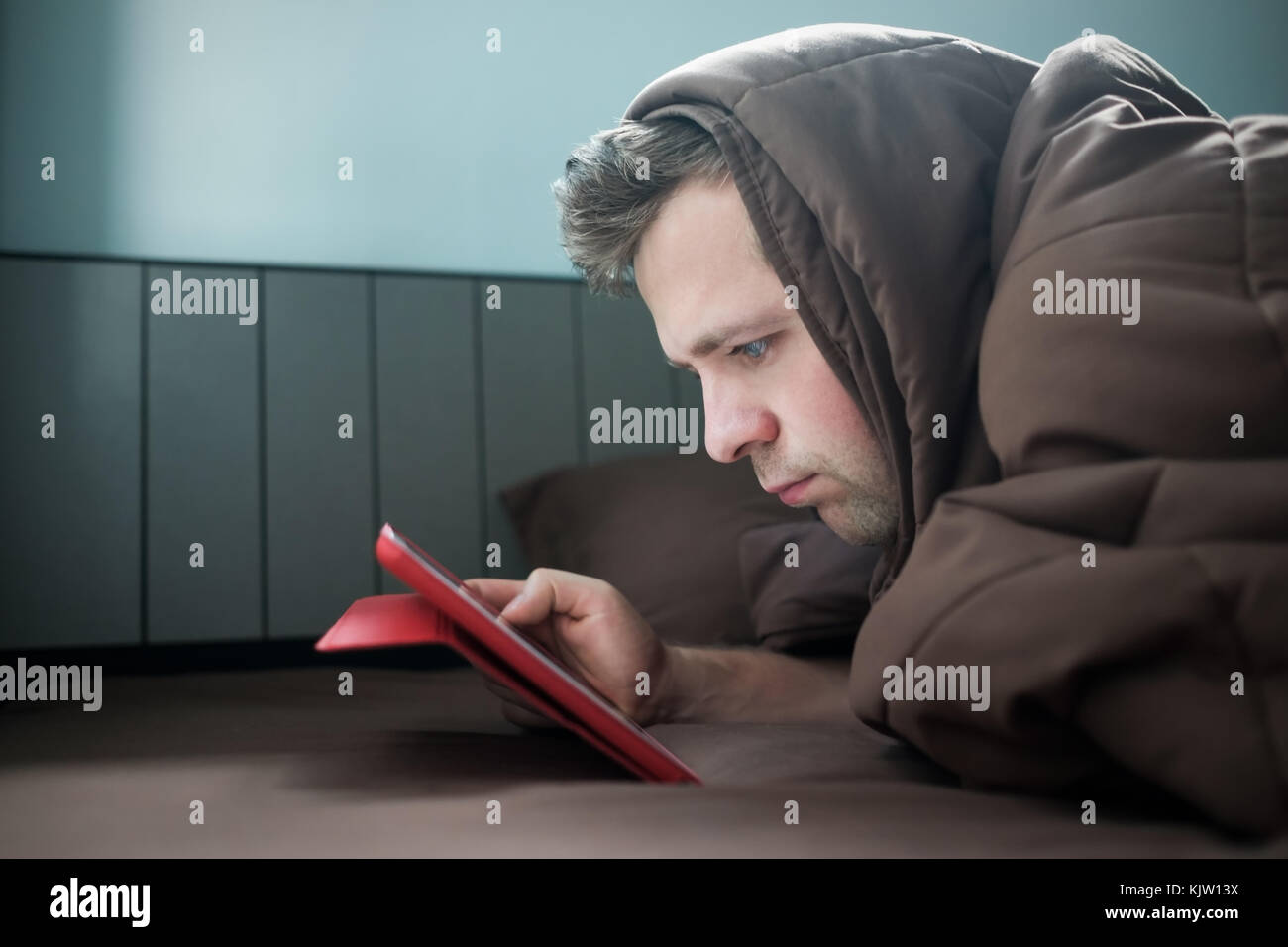 Man is working at night, lying on the sofa in the living room with tablet, covering his head with blanket. Stock Photo