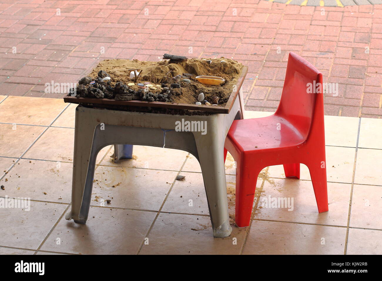 A child's small sand pit on a small table and a small red chair with sand, stones, lids and shells in landscape format with copy space Stock Photo