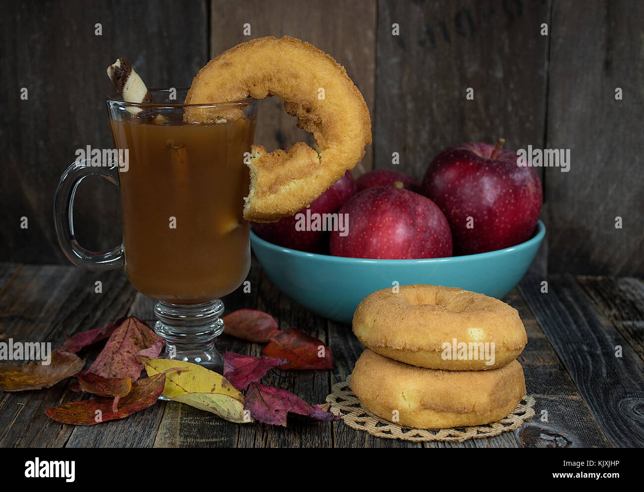 donut on edge of apple cider in glass mug with fall leaves and red apples in turquoise bowl on rustic wood Stock Photo