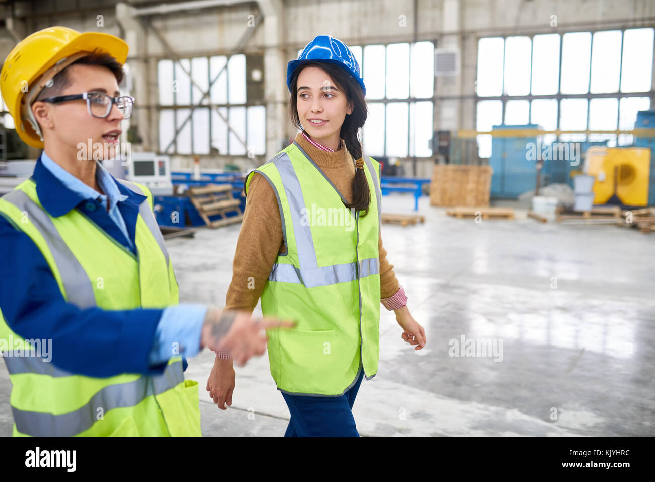 Confident experienced forewoman walking with female colleague over factory workshop and explaining her work Stock Photo