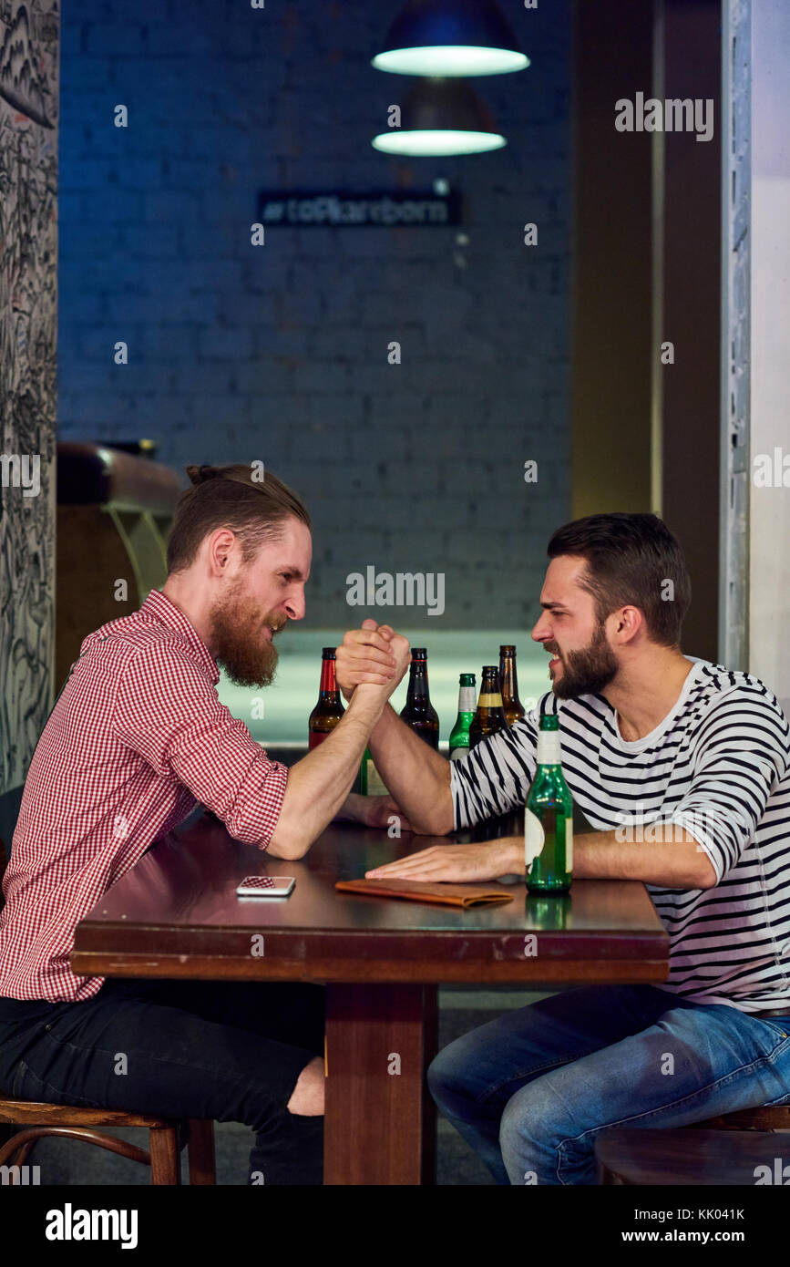 Side view portrait of two modern young men armwrestling sitting at table in bar and drinking beer, copy space Stock Photo