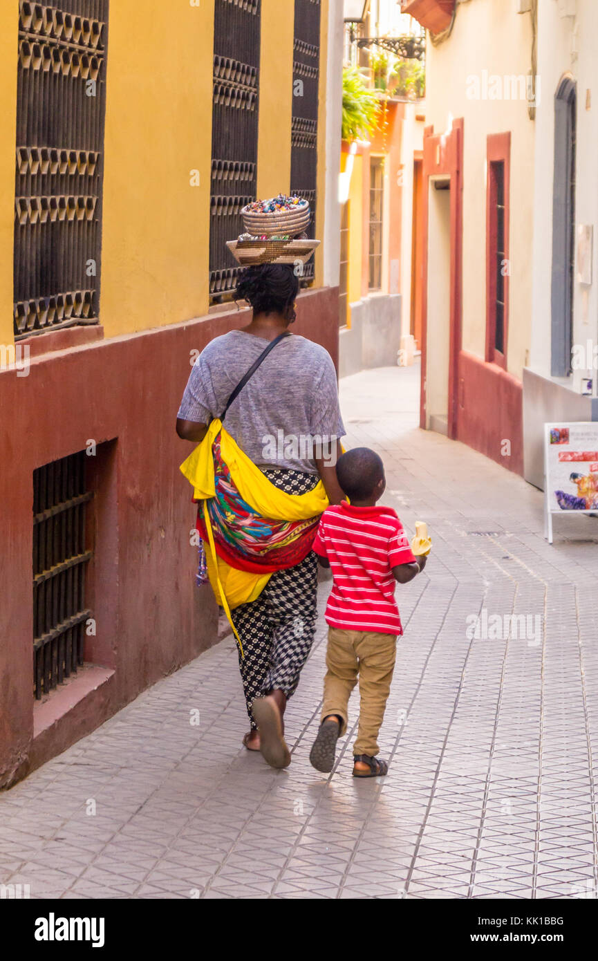 African trinket seller carrying her wares in a basket on her head, and her son in a back street, Seville, Andalucia, Spain. Child eating a banana Stock Photo