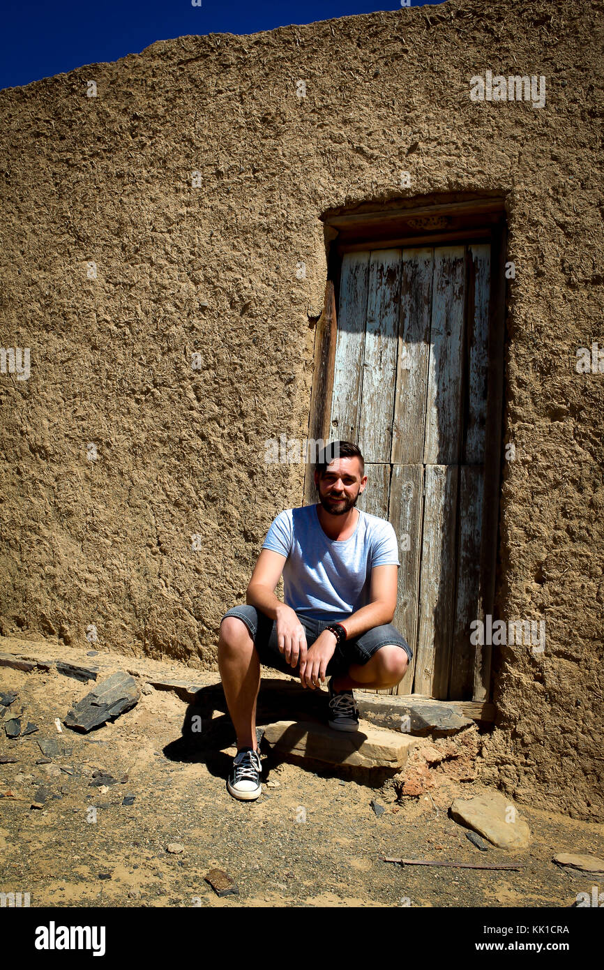 Caucasian man wearing t-shirt and jean shorts crouching in front of old door of mud house Stock Photo