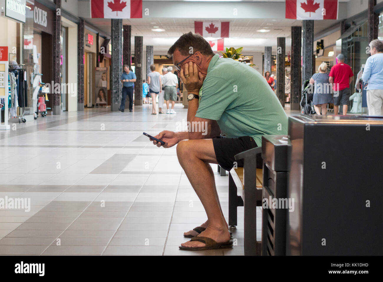 Man sits on bench in shopping mall checking his cell phone in Lindsay