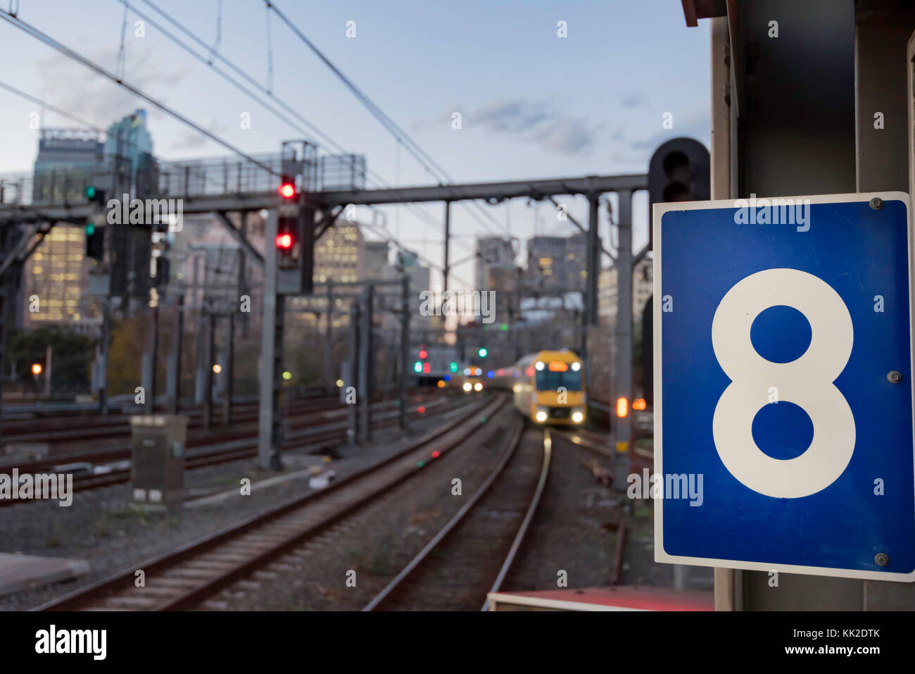 Prominent number 8 sign at a railway station with blurred train tracks, the city and a train approaching in the background at Central Station, Sydney Stock Photo