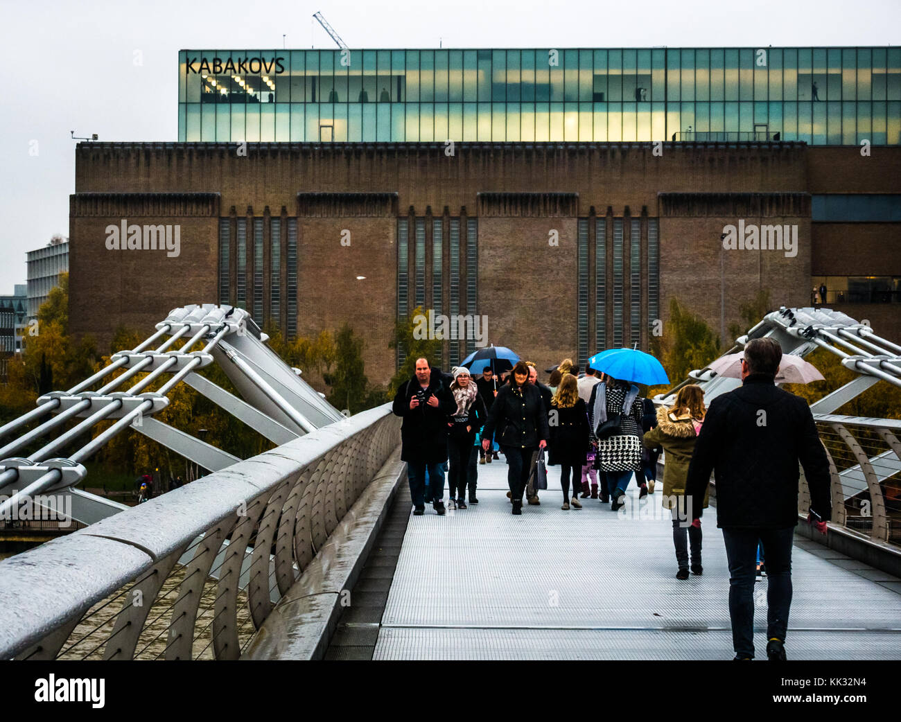 People walking on Millennium Bridge on rainy day with umbrellas to lit up Tate Modern Art Gallery with Kabakovs exhibition, London, England, UK Stock Photo