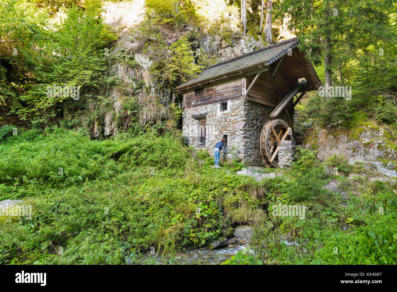 Alpine landscape with old water mill in the forest thicket. Woman tourist peek into window. Western Carinthia, Austria. Stock Photo