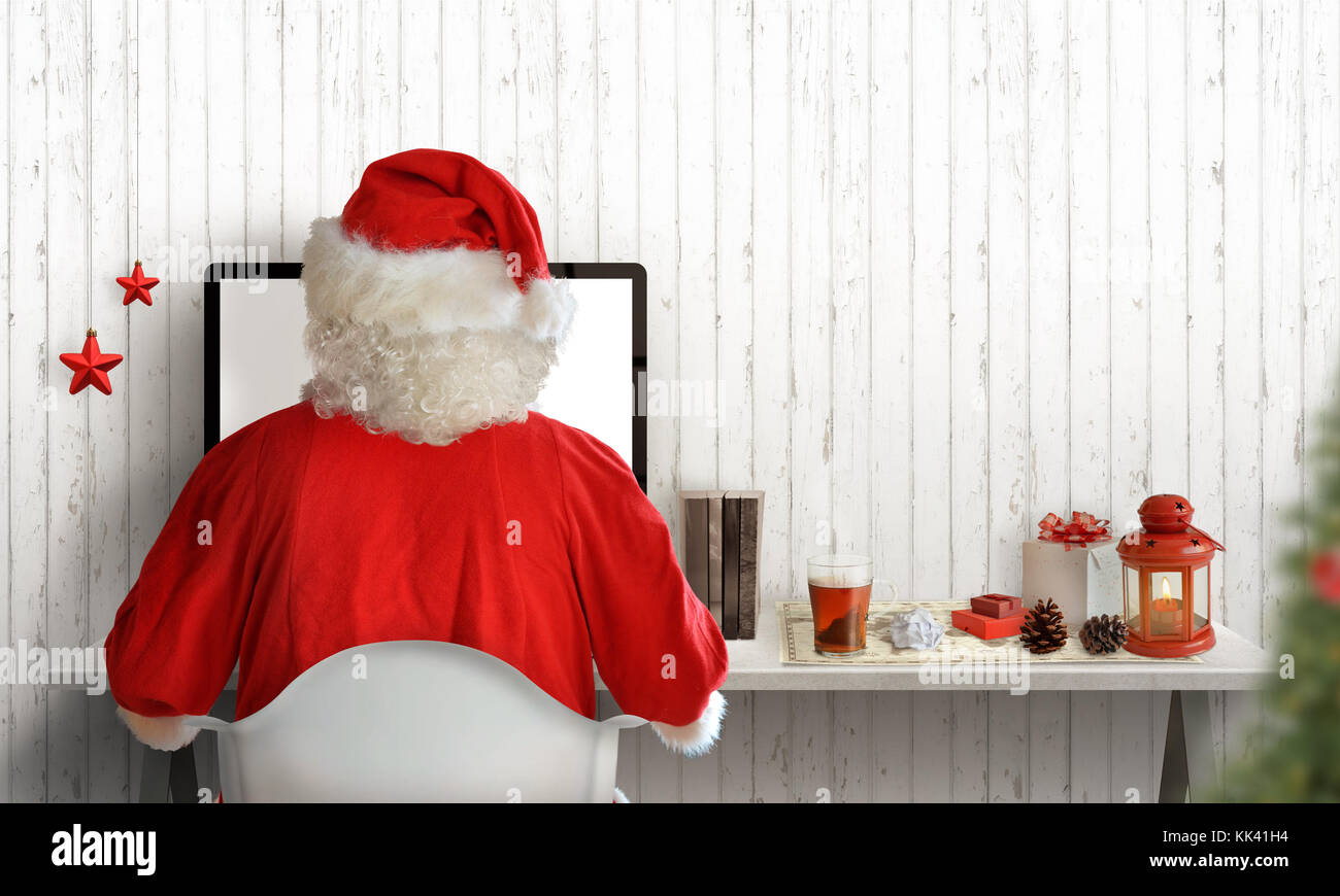 Santa Claus work on computer and send letters to kids. Christmas tree, gifts, and decorations beside. Free space for greeting text. Stock Photo