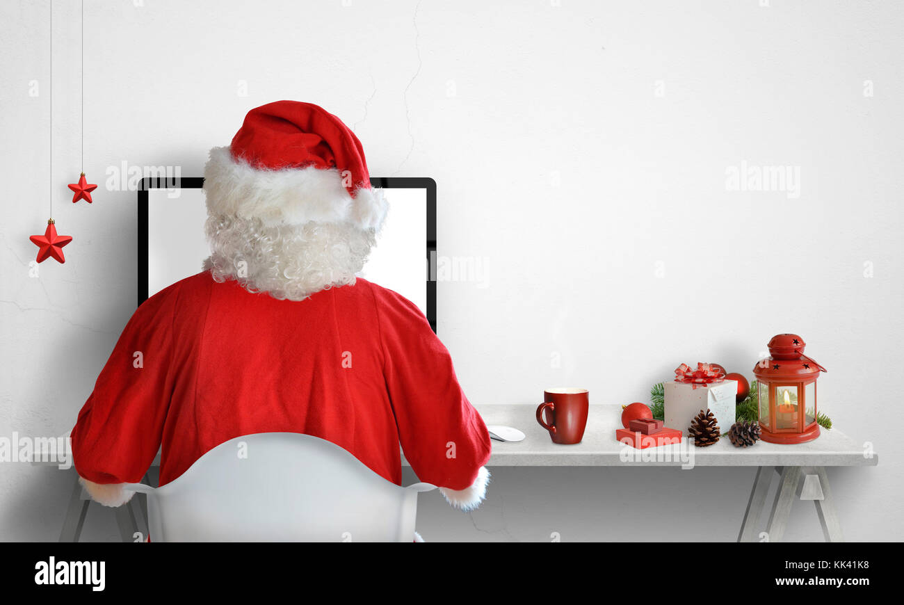 Santa Claus work on computer. Christmas decorations beside with free space on the wall for text. Stock Photo