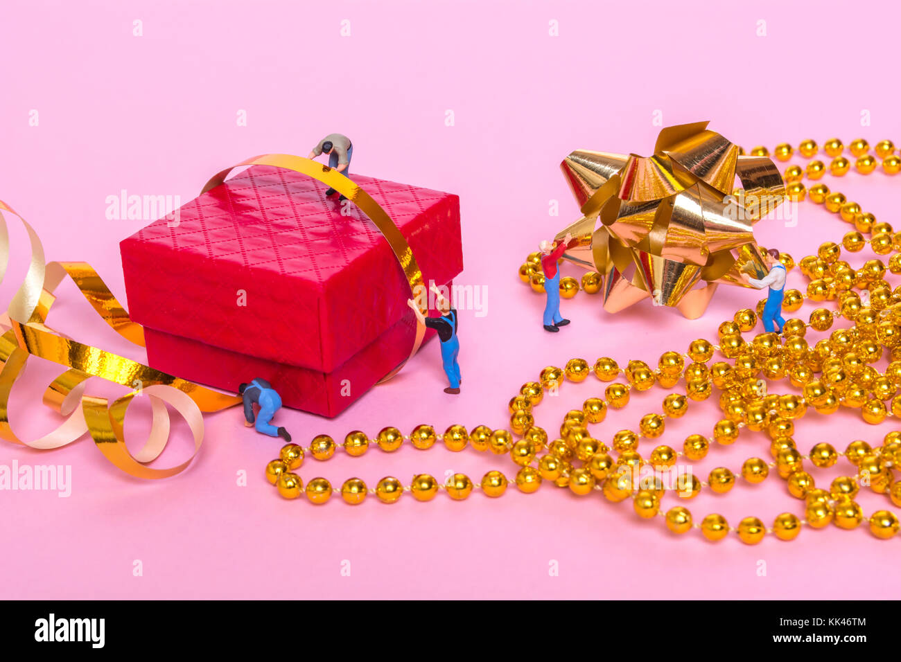 Creative concept with Miniature people and a gift box on a pink background. The process of packing gifts. Gold ribbon, bow, festive beads. Creative co Stock Photo