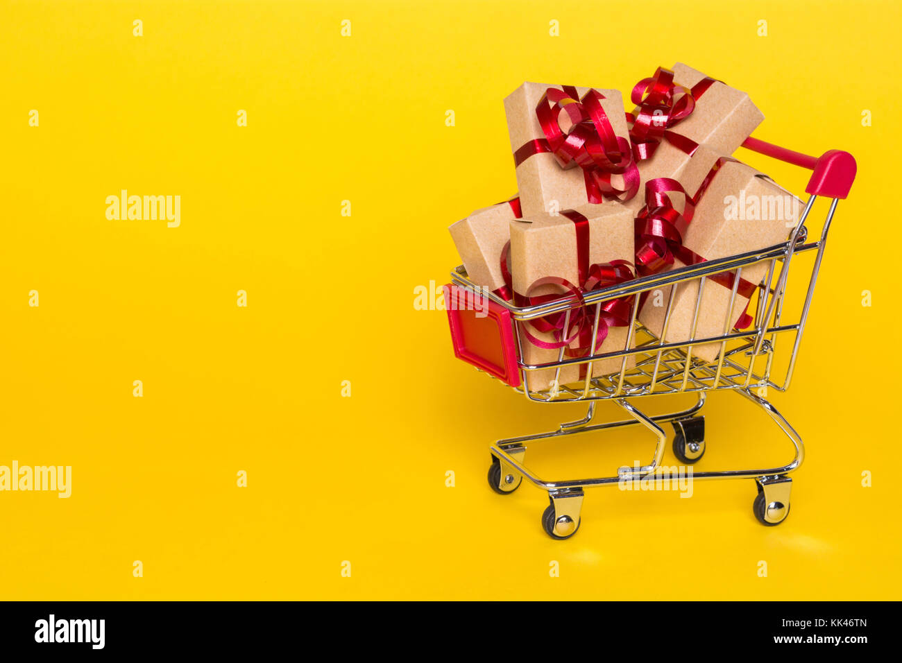 Creative concept with shopping trolley with gifts on a yellow background. Gifts wrapped in kraft paper with a red ribbon and bow. Stock Photo