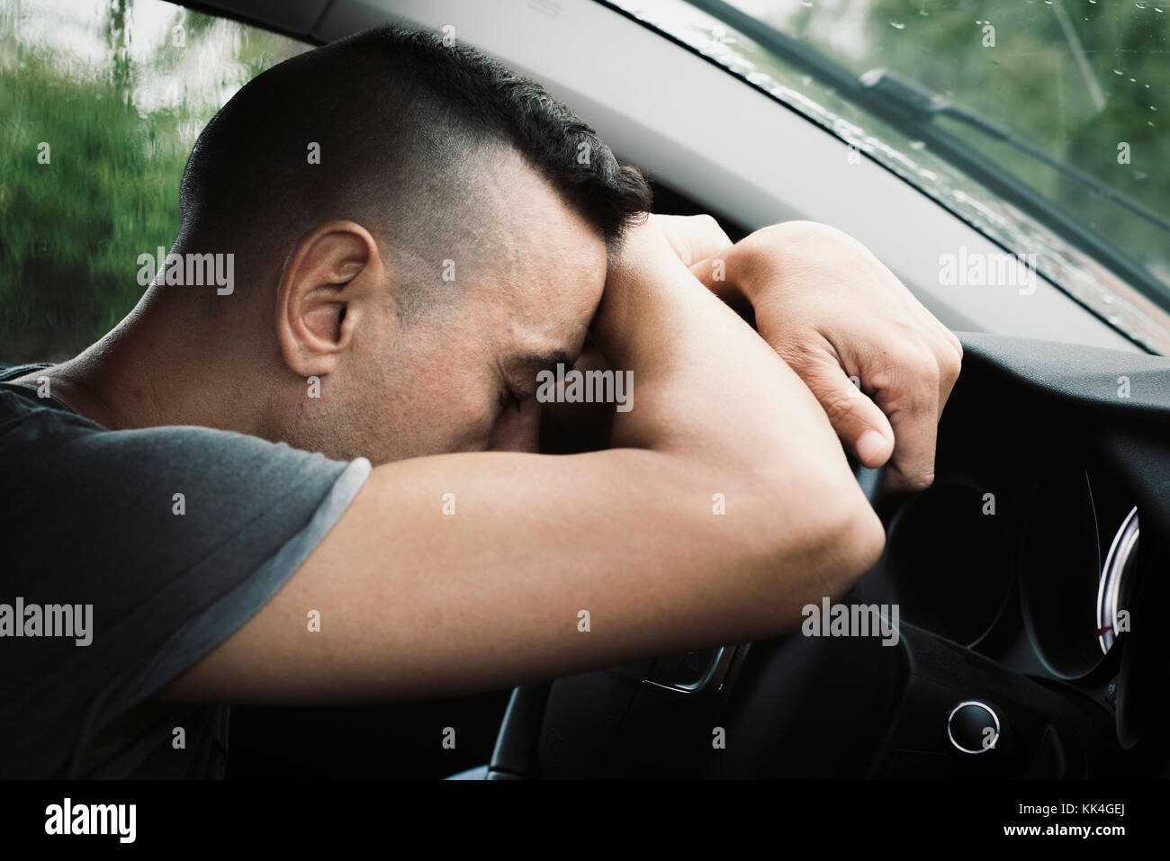 closeup of a young caucasian man sitting at the driver seat of a car reclining his head on the steering wheel, in a rainy day Stock Photo
