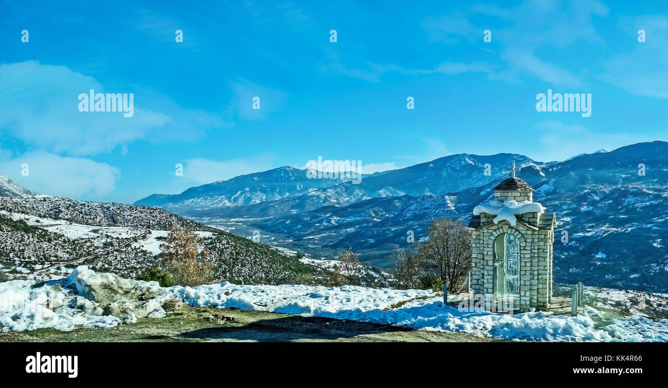 The tiny roadside chapel, covered with snow, with the amazing landscape of Pindus mountains on the background, Greece. Stock Photo
