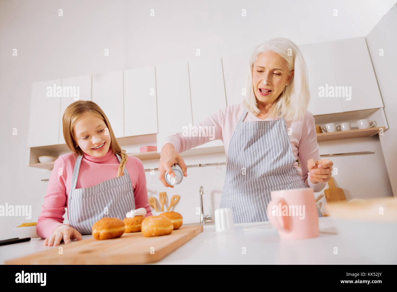 Favourite hobby. Cute happy delighted grandmother and granddaughter standing in the kitchen and putting whipped cream on buns while enjoying cooking Stock Photo