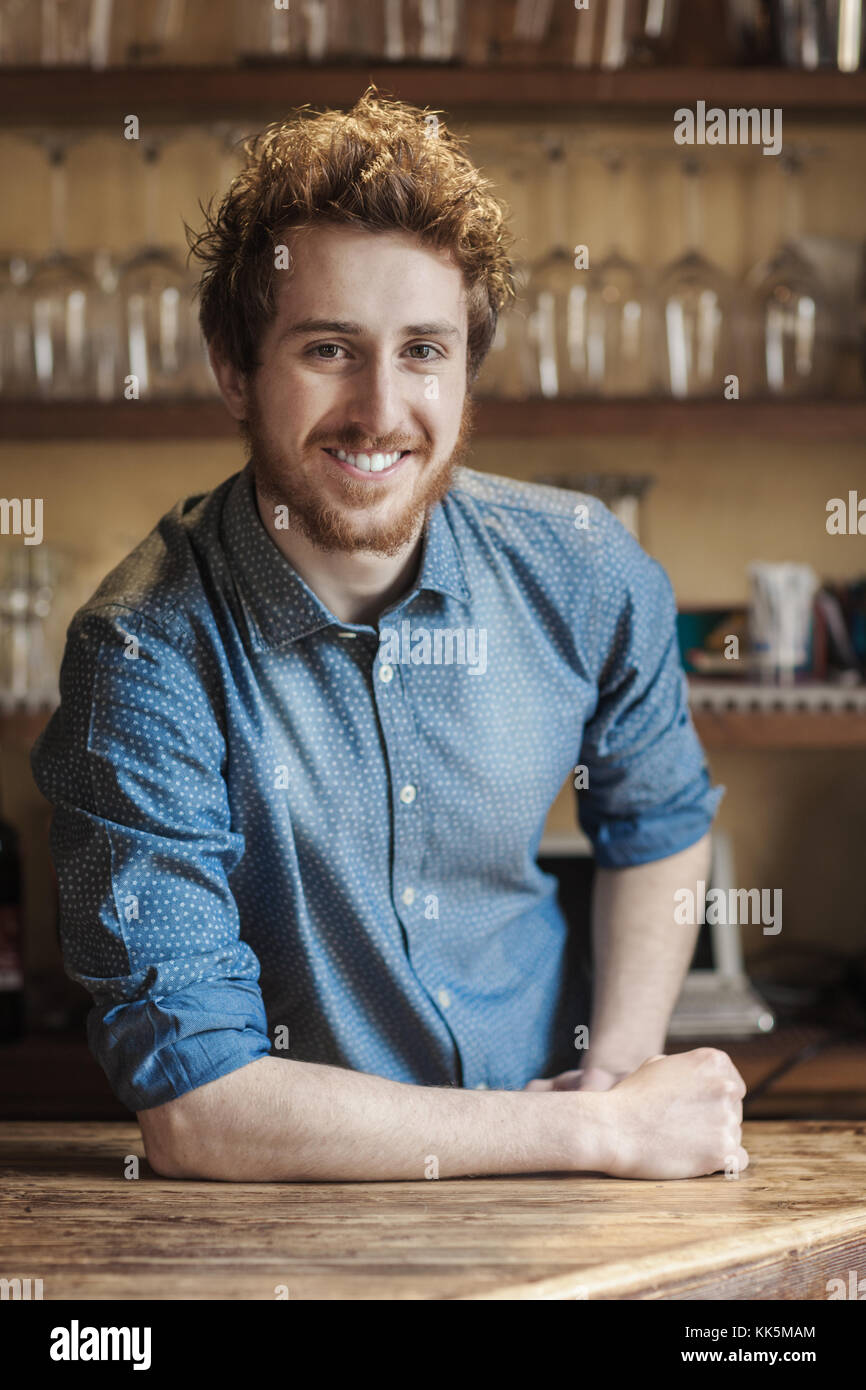 Young barman leaning on wooden bar counter and smiling at camera, shelves with glasses on background Stock Photo