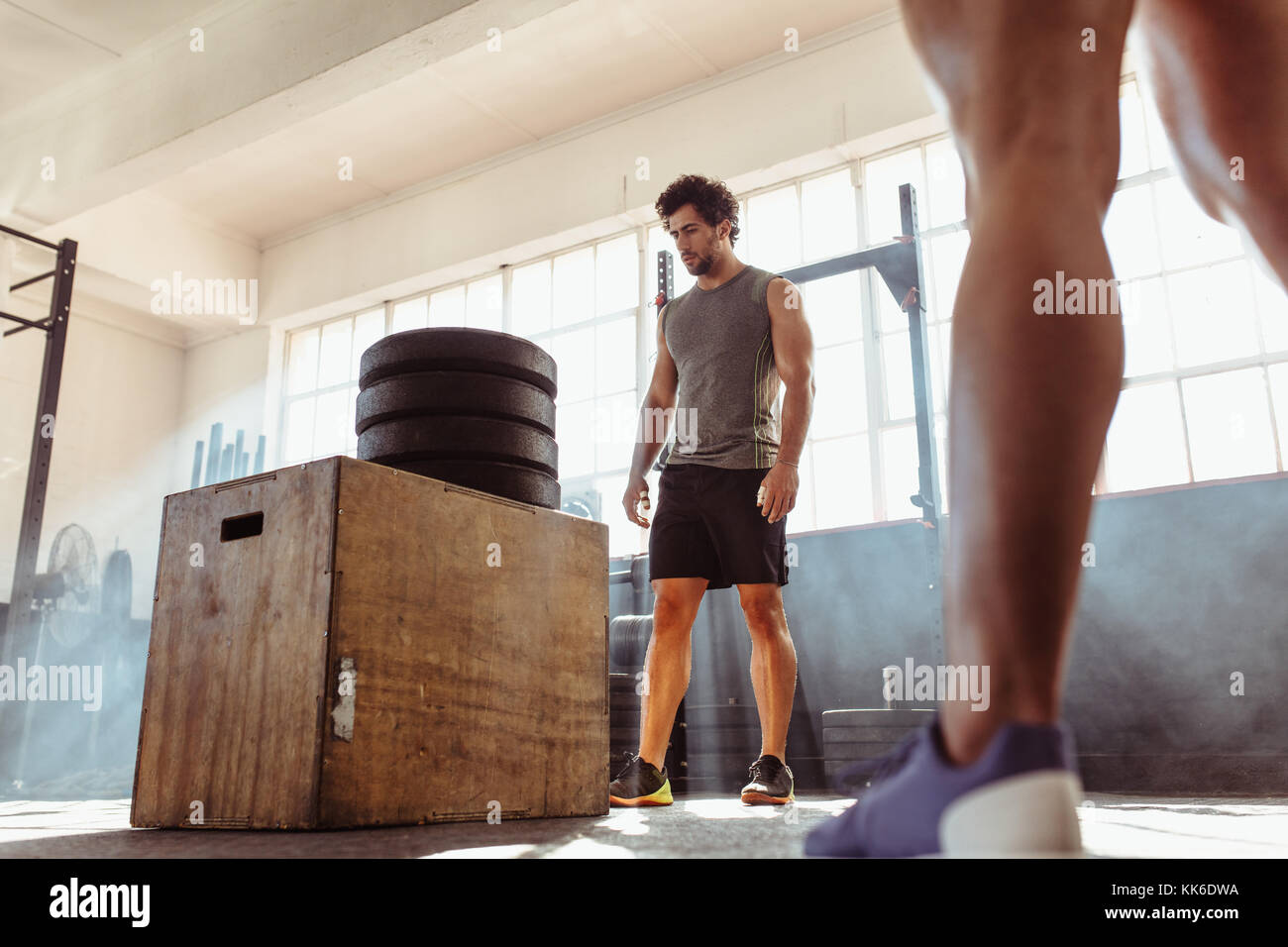 Determined fit man doing box jumping in health club. Muscular male athlete standing by wooden box with extra height made by pile of weights. Stock Photo