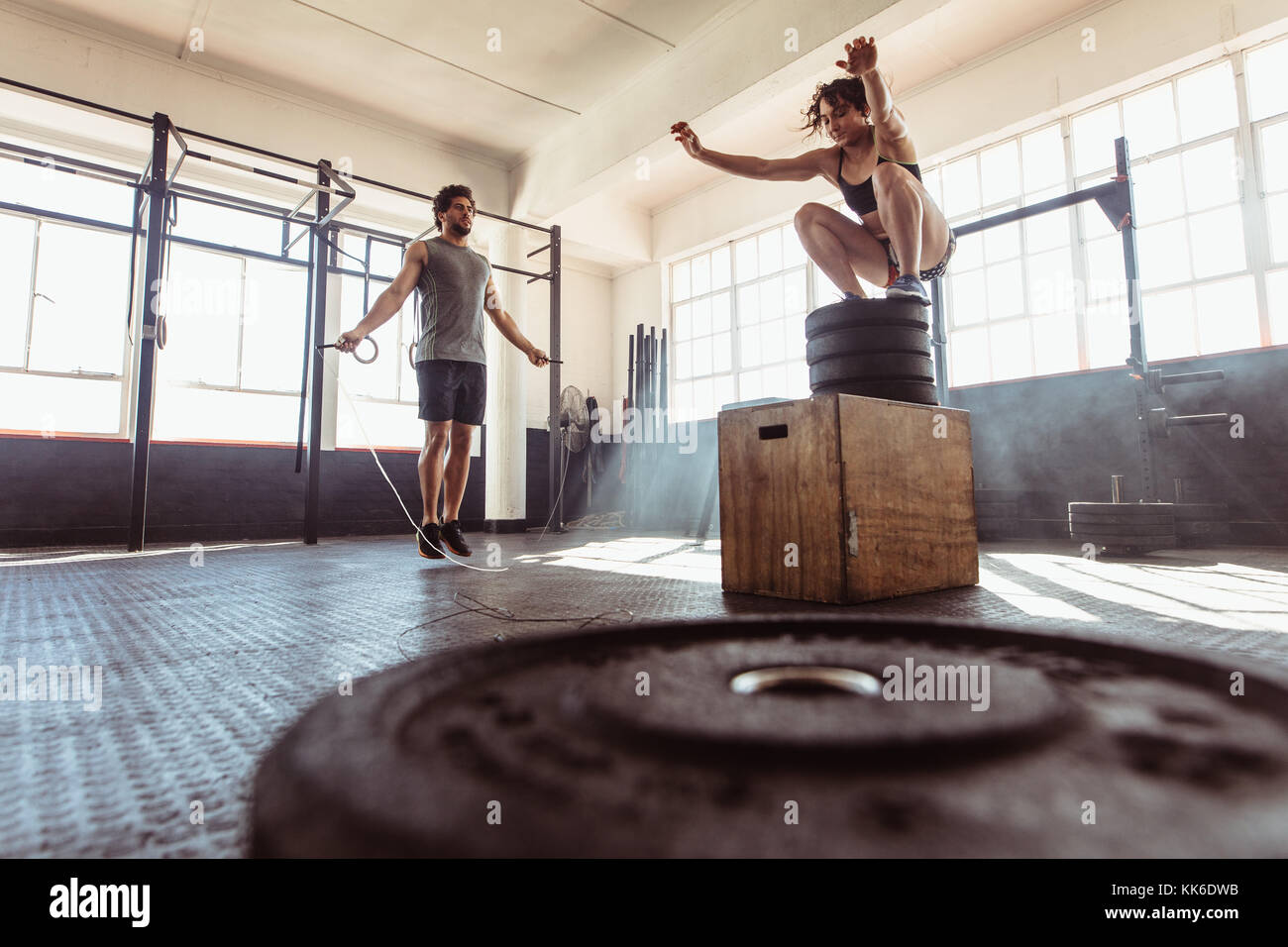 Man exercising using skipping rope and woman jumping on box in gym. Athletic couple training hard at the cross fit gym. Stock Photo