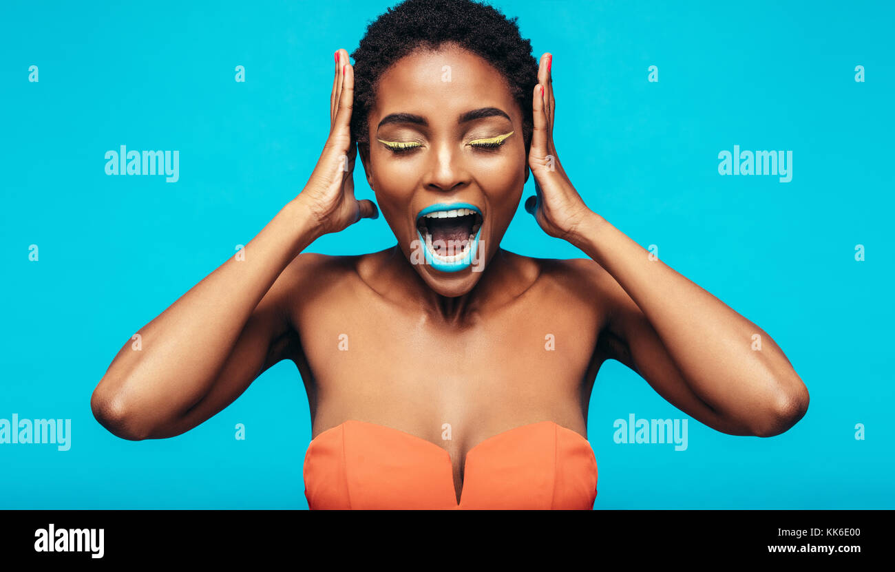 African woman with colorful makeup screaming against blue background. Female fashion model with mouth open and hands on ears. Stock Photo