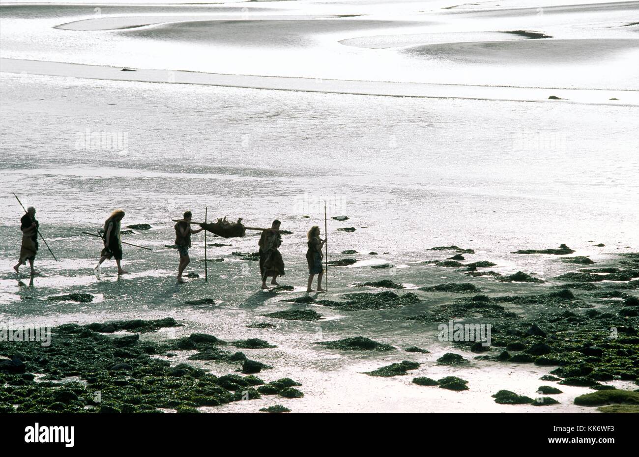 Neolithic Mesolithic prehistoric caveman cavemen cave man hunter gatherer walking on mud flat tidal estuary carrying deer food carcass and spears Stock Photo
