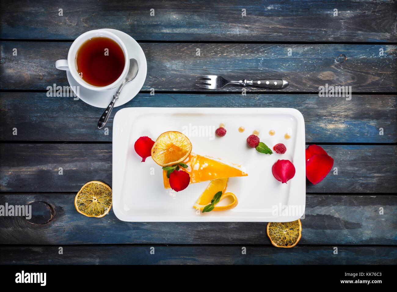 Orange cake with a cup of tea fresh raspberries on a white plate with rose petals. Top view. Beautiful wooden background. Stock Photo