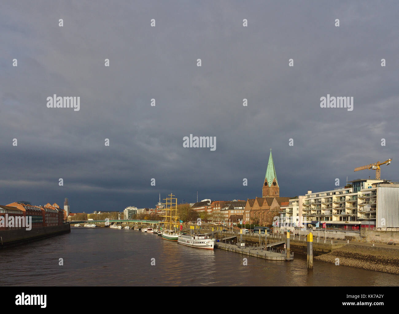 Riverside view of Bremen, Germany with moored ships, historic church, residential buildings and dark rain clouds in the background Stock Photo