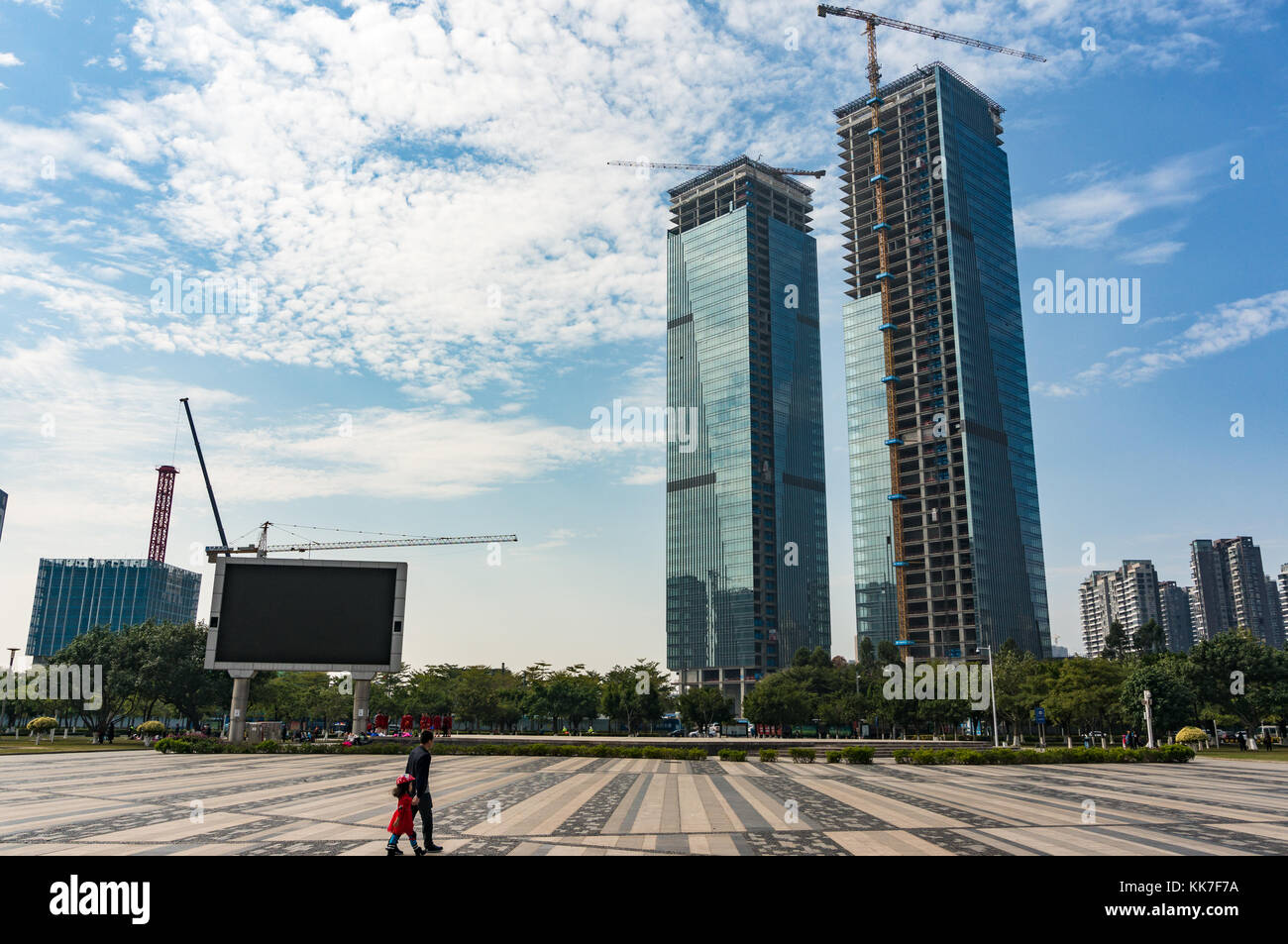 Father and daughter walking in vast city square as unfinished skyscrapers buildings loom in the distance in Shenzhen, Guangdong Province, China Stock Photo