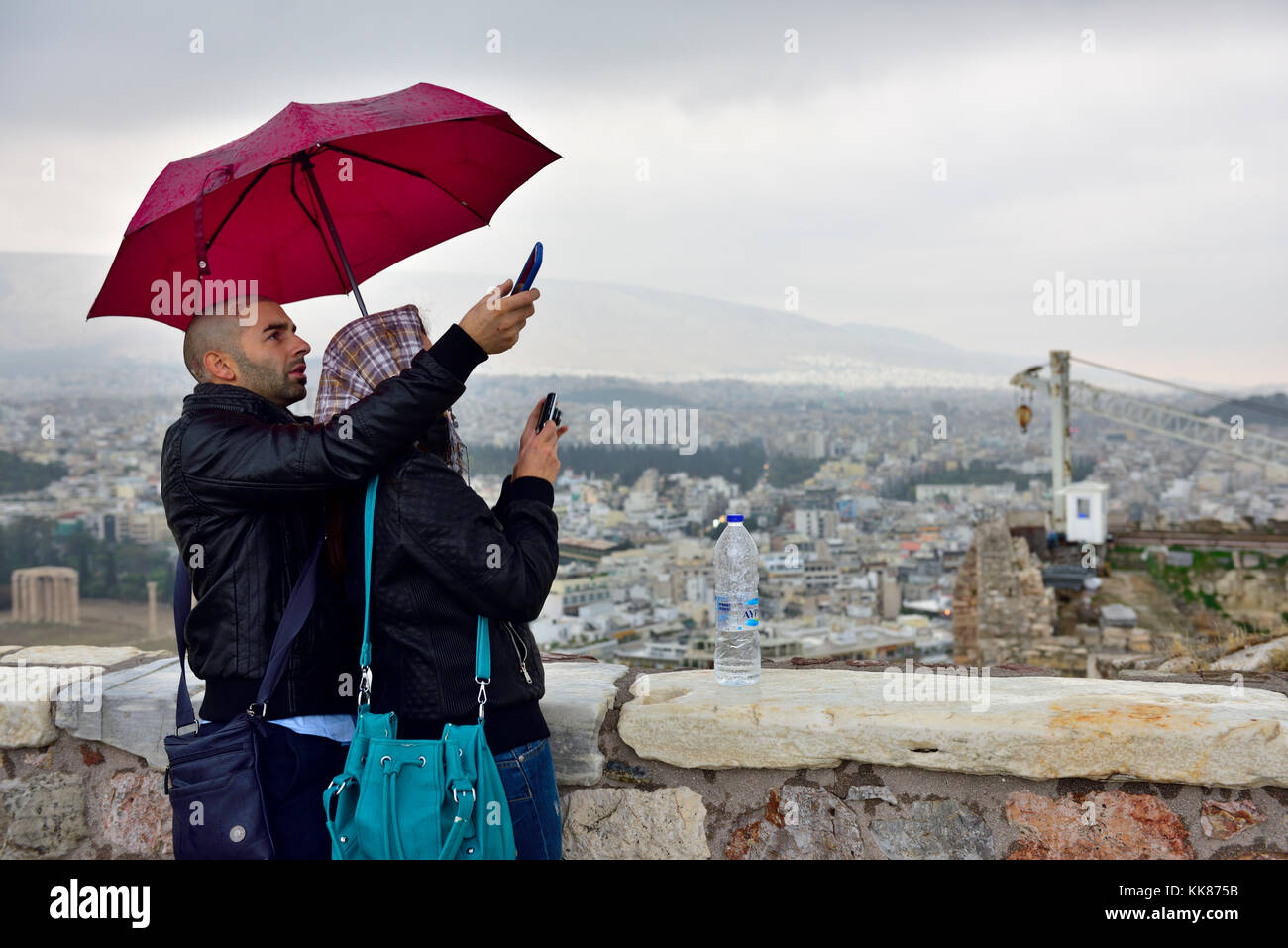 Couple with umbrellas in rain with mobile phones taking photos overlooking Athens from the Acropolis Stock Photo