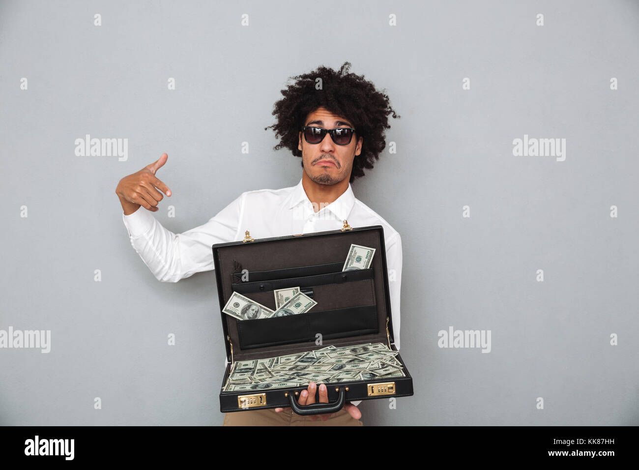 Portrait of a young afro american man in sunglasses pointing finger at briefcase full of money banknotes isolated over gray background Stock Photo
