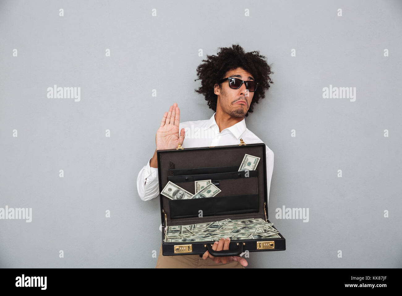 Portrait of a funny young afro american man in sunglasses holding briefcase full of money banknotes isolated over gray background Stock Photo
