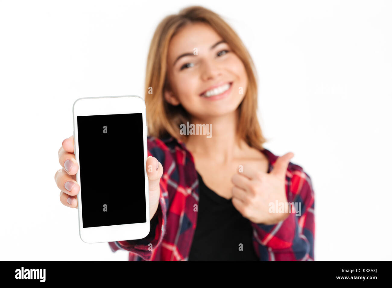 Close up portrait of a smiling cheerful girl showing blank screen mobile phone and giving thumbs up isolated over white background, focus on mobile ph Stock Photo