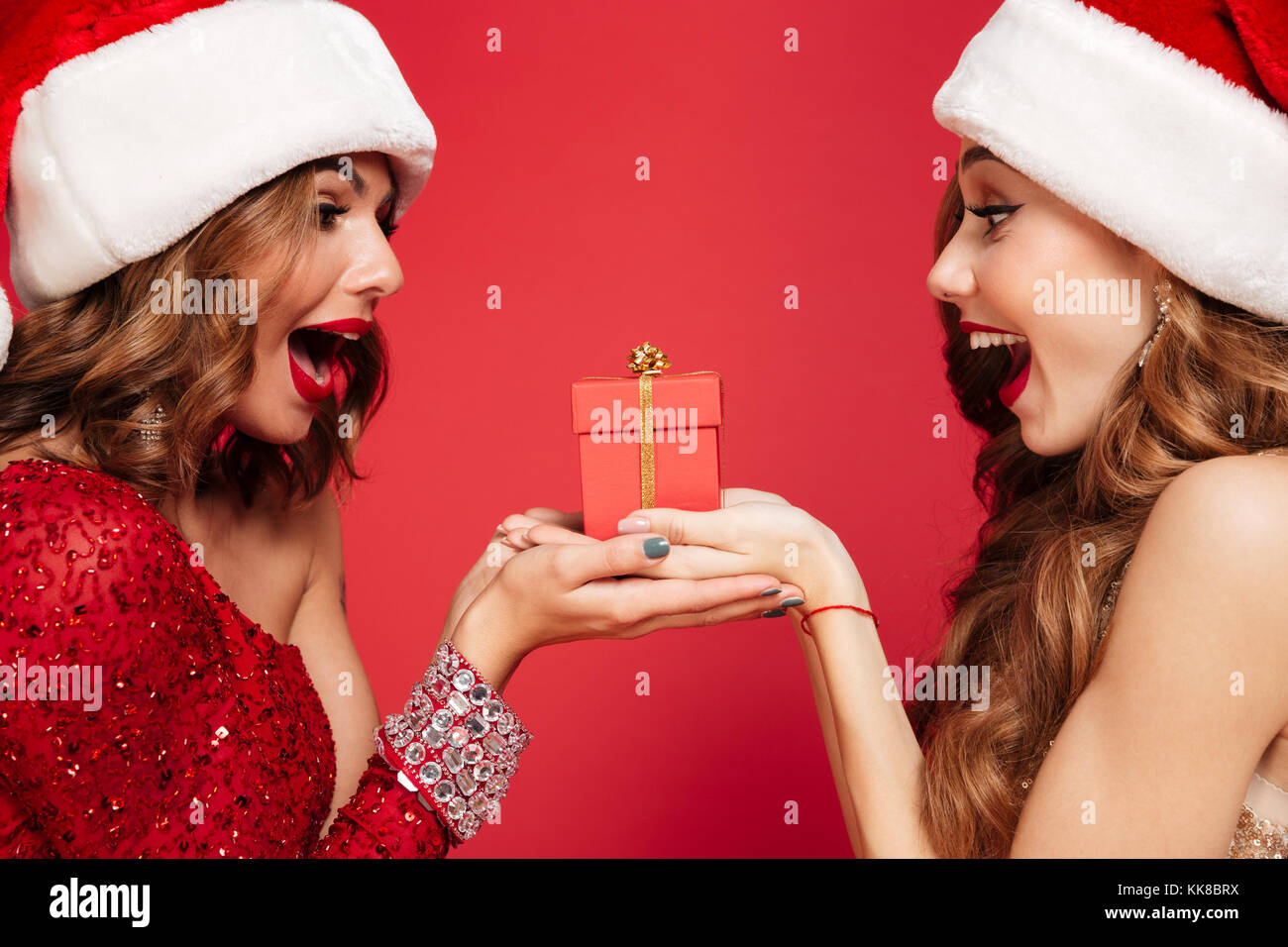 Close up portrait of two cheerful excited women in christmas hats holding a gift box and looking at each other isolated over red background Stock Photo