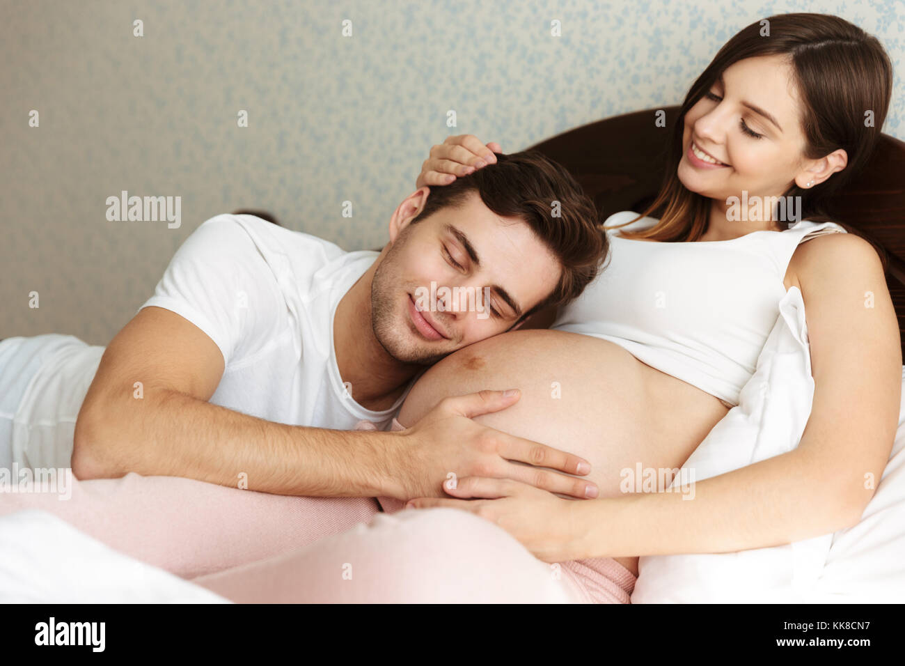 Smiling young pregnant wife lying in bed with her husband in the bedroom, man is lying on her belly Stock Photo