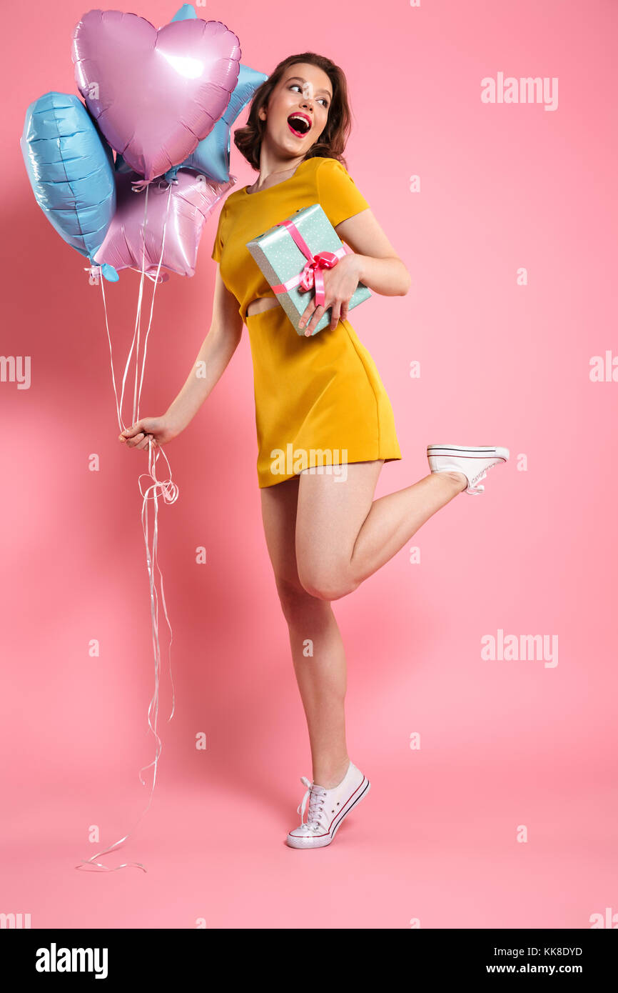 Image of excited cheerful young lady in yellow dress posing isolated pink background and holding ballons with gift box. Looking aside. Stock Photo