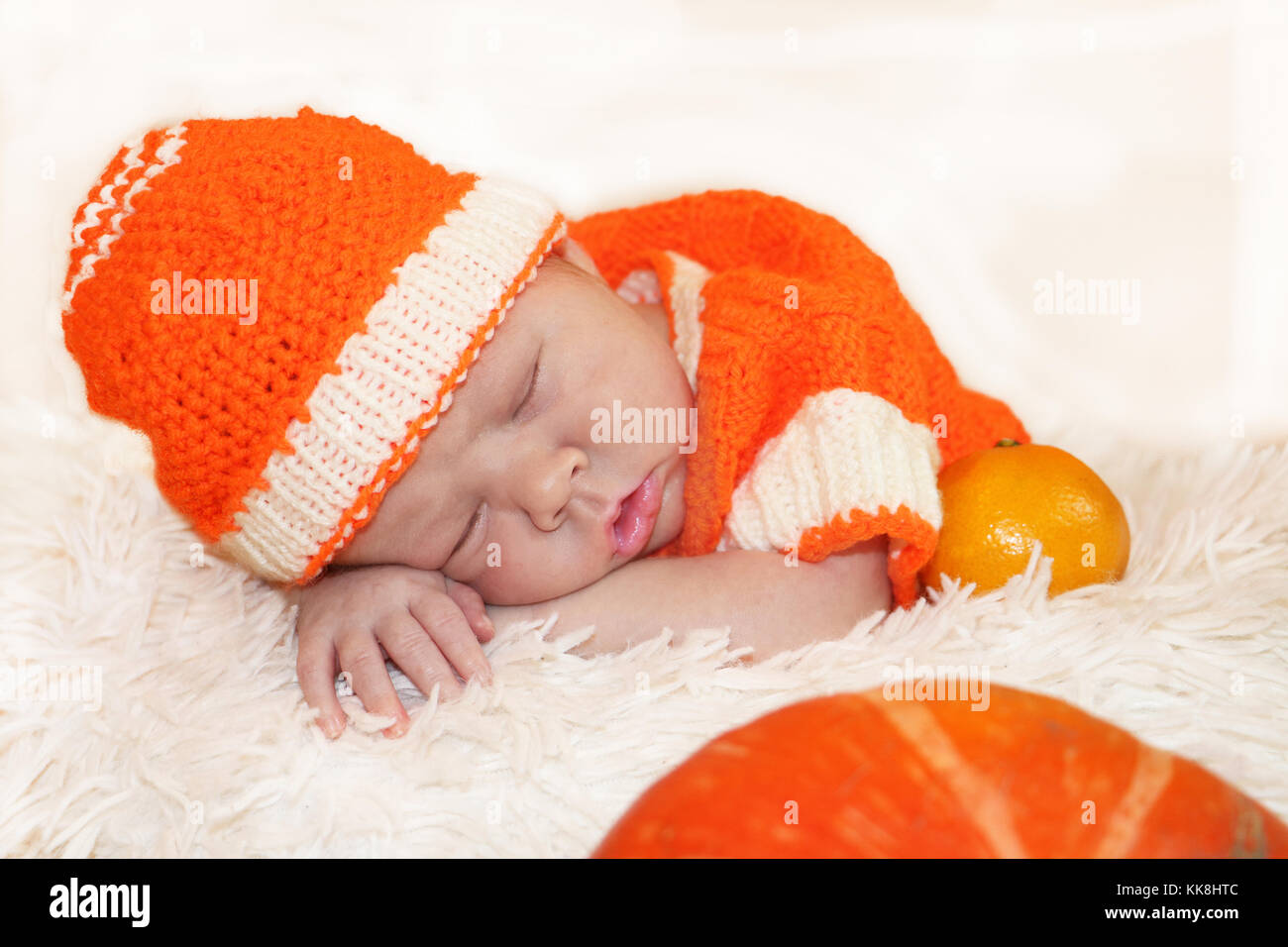 Newborn baby in knitted orange costume sleeping on white blanket with pumpkin and oranges in