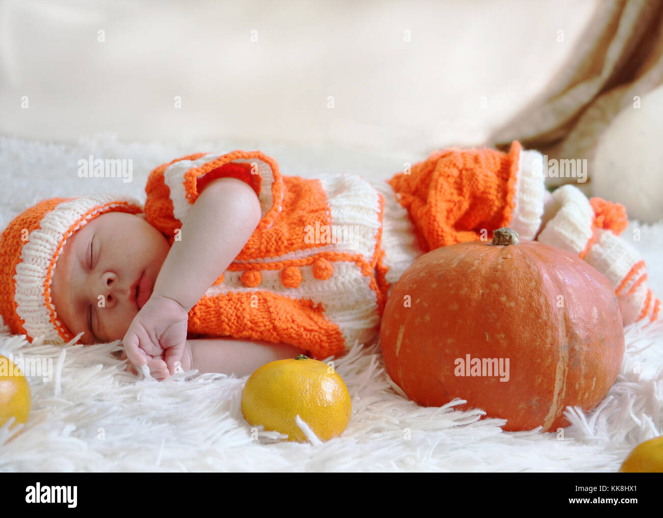 Newborn baby in knitted orange costume sleeping on white blanket with pumpkin and oranges in front of him. Halloween or Thanksgiving concept. Stock Photo