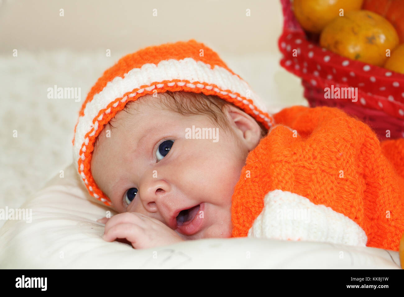 Astonished cute newborn baby dressed in a knitted orange costume with  oranges in basket behind of him. Autumn halloween or harvest concept. Baby face Stock Photo