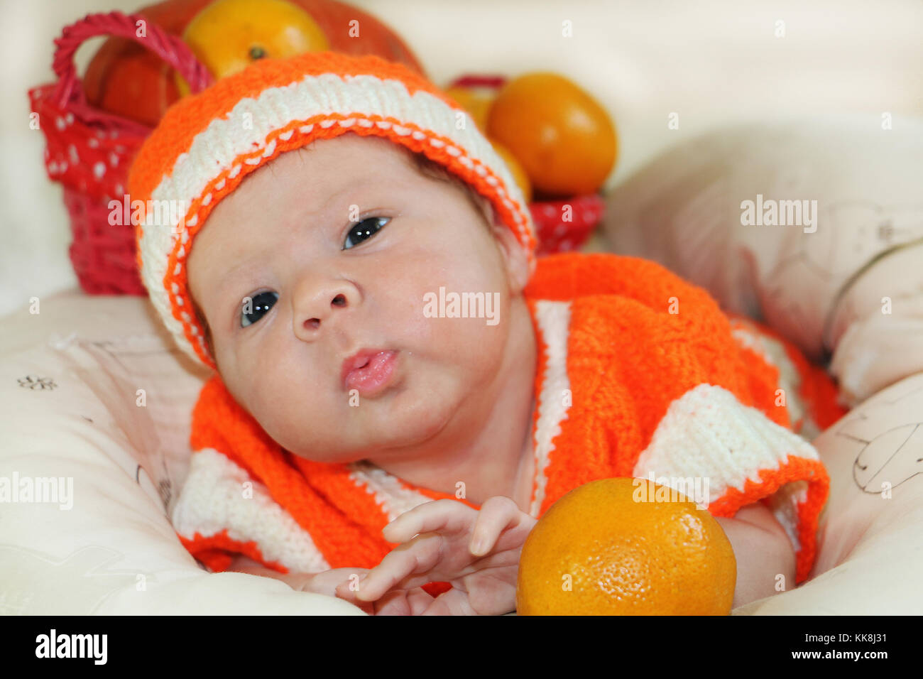 Baby sending air kiss. Portrait of newborn baby in orange knitted costume on white blanket and basket with oranges and pumpkin behind of him sending a Stock Photo
