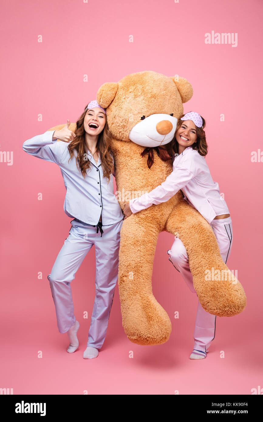 Photo of two amazing friends women in pajamas isolated over pink background hug big teddy toy bear. Looking camera showing thumbs up Stock Photo Alamy