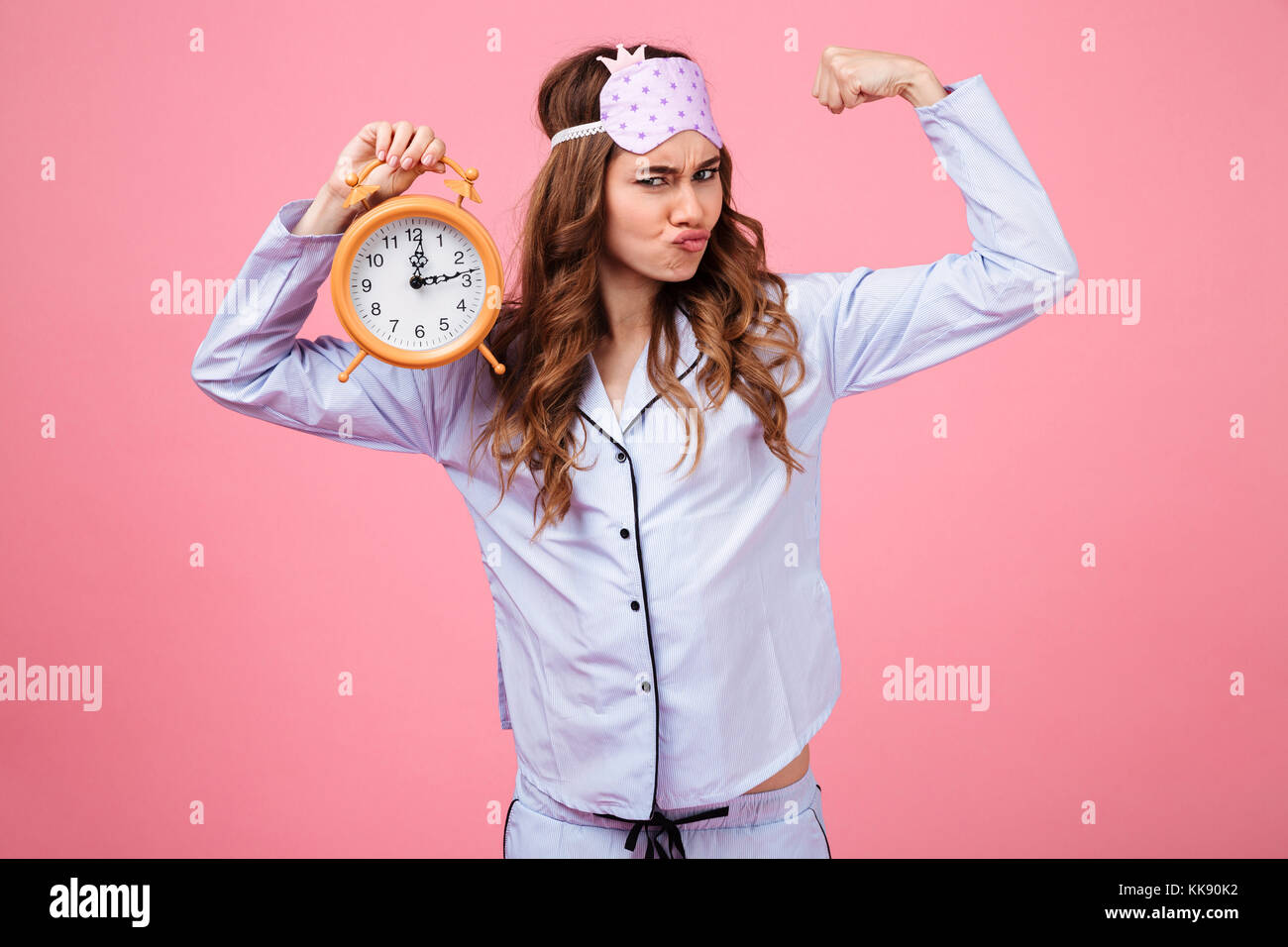Portrait of a funny young girl dressed in pajamas holding alarm clock and flexing biceps isolated over pink background Stock Photo